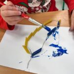 Dziecko maluje niebieską farbą korale zrobione z nawleczonego na nitkę makaronu