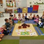 Dzieci siedzą w sali przedszkolnej - pozują do zdjęcia ze zrobionymi z dłoni "serduszkami". Pośrodku nich na dywanie leżą wykonane prace plastyczne